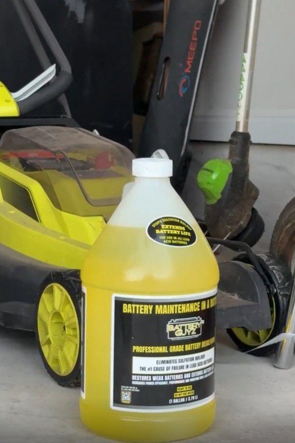 A product bottle on the ground next to a lawn mower for size context. Caption: Scale example 1. Product shown near everyday objects for size context.